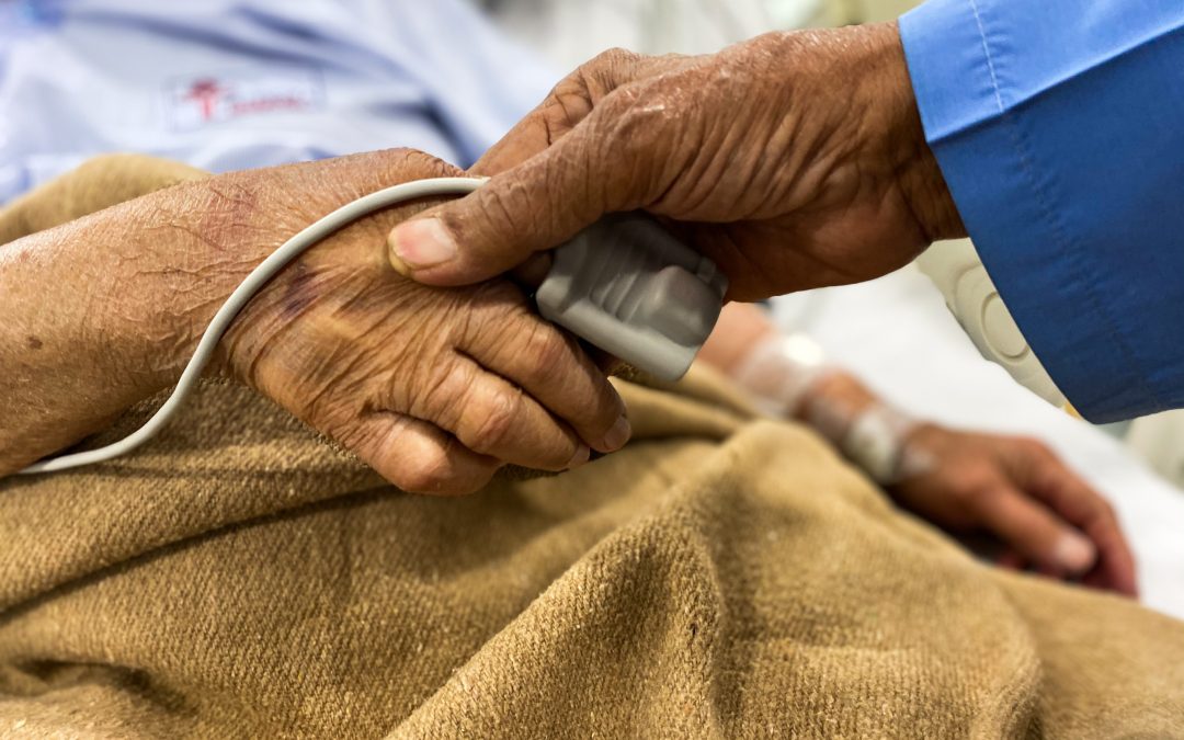 An elderly couple hold hands. One is lying in a hospital bed with an oxygen meter on their finger.
